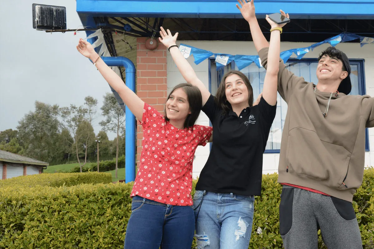 Fotografía de dos jóvenes alumnas y un alumno del Cibercolegio en el Campus de la Fundación Universitaria Católica del Norte, se encuentran alzando los brazos y están en la zona verde disfrutando del colegio. Una de las alumnas es la personera estudiantil electa en el año 2022.