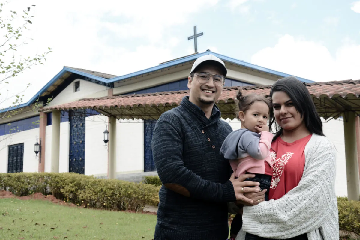 Fotografía de una familia conformada por padre, madre de hija. Ellos están en el campus de la Fundación Universitaria Católica del Norte y en el fondo se observa la capilla.