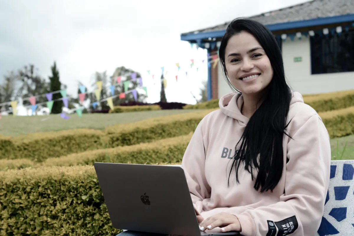 Fotografía de madre de familia en el Campus de la Fundación Universitaria Católica del Norte, en Santa Rosa de Osos Antioquia, Colombia. En la imagen se puede ver a la madre de familia sosteniendo un portátil, en el que se dispone a realizar una solicitud en la secretaria académica a través de la plataforma de padres. Al fondo se observa el hermoso paisaje verde del campus universitario.
