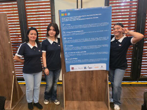 La docente acompañante y dos estudiantes del Cibercolegio UCN posan junto al póster del proyecto ECCpoint durante un evento académico sobre sostenibilidad. El panel presenta información sobre una propuesta de economía circular en la ruralidad, incluyendo el reto abordado, la pregunta de investigación, la ruta de trabajo, la idea central y los aprendizajes obtenidos. La exhibición se desarrolla en un espacio de presentación de proyectos con paneles informativos y ventanales al fondo.