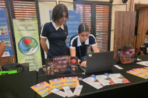 Dos estudiantes del Cibercolegio UCN presentan un proyecto en un stand informativo durante un evento académico sobre sostenibilidad. Sobre una mesa negra se observan un computador portátil, folletos, material promocional y una pantalla con información institucional. Al fondo hay afiches relacionados con investigación ambiental y cuidado del planeta, entre ellos un pendón del semillero de investigación “Amor por el Planeta”. Las estudiantes interactúan con el computador mientras exponen su iniciativa en un espacio de intercambio de proyectos y experiencias ambientales.